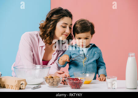 Felice madre guardando carino piccolo figlio di uova di mantecazione su sfondo bicolore Foto Stock