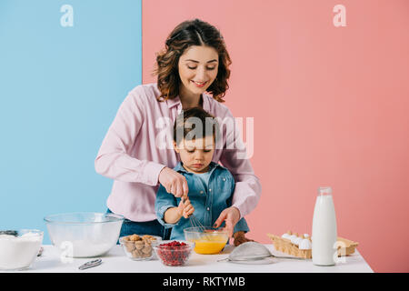 Sorridente madre aiutare piccolo figlio in uova di mantecazione con palloncino frusta su sfondo bicolore Foto Stock