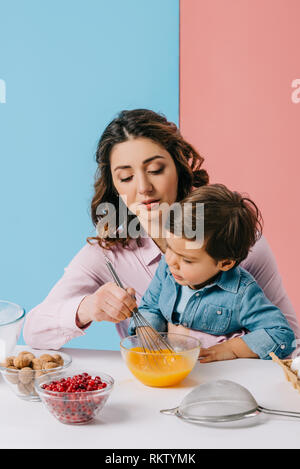 Madre seduta con piccolo figlio da tavolo da cucina e monta le uova in una terrina su sfondo bicolore Foto Stock