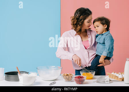 Azienda madre piccolo figlio mentre le uova di mantecazione con palloncino frusta su sfondo bicolore Foto Stock