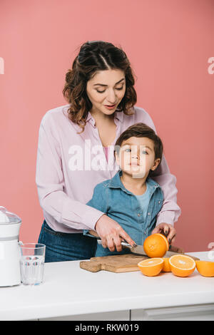 Felice madre con carino piccolo figlio di taglio di arance fresche insieme isolato in rosa Foto Stock