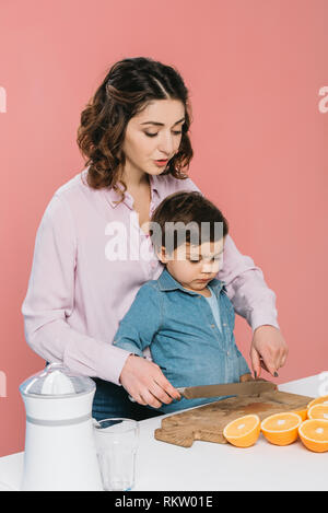 La madre mostra tagliare le arance al piccolo figlio mentre si tiene il coltello isolato in rosa Foto Stock