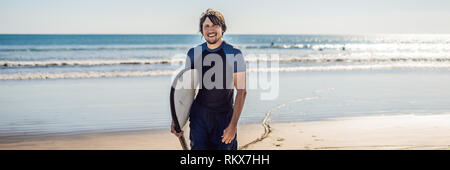 Bello giovane sportivo surfer in posa con la sua tavola da surf sotto il suo braccio nella sua muta su una sabbiosa spiaggia tropicale banner formato lungo