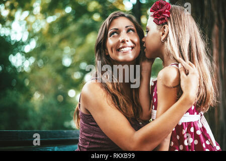 Bella madre e figlia sono divertirsi al parco nella giornata di primavera. Bambina è sussurra qualcosa a sua madre. Foto Stock