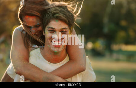 In prossimità di un paio di divertirsi giocando holi. Uomo sorridente portando la sua fidanzata sulla schiena durante la riproduzione di holi all'esterno. Foto Stock