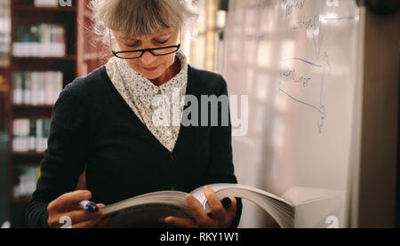 In prossimità di una donna anziana in piedi in una libreria capovolgimento di pagine di un libro di testo. Donna senior per il controllo dei libri di riferimento in una biblioteca universitaria. Foto Stock