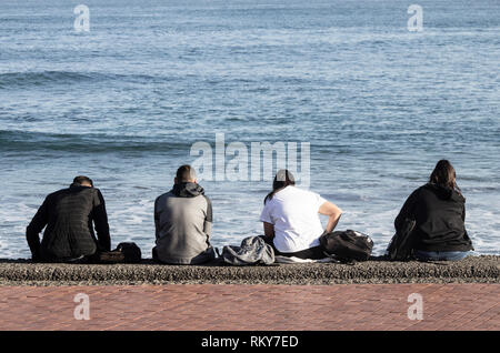 Vista posteriore di quattro adolescenti seduti sulla parete del mare e guardando i telefoni cellulari: la noia, annoiato adolescente, social media... concetto. Foto Stock