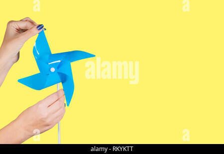 Primo piano delle mani femminili con manicure blu blu tenuta girandola isolati su giallo brillante con sfondo copyspace. Orizzontale fotografia a colori Foto Stock