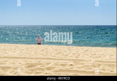 Donna matura in una camicia bianca e cappello bianco è in piedi sulla spiaggia e guardare il mare nella soleggiata giornata estiva. Foto Stock