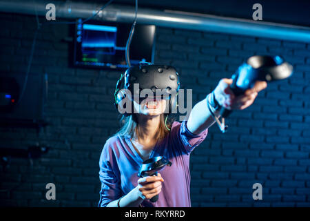 Giovane donna un gioco utilizzando la realtà virtuale auricolare e gamepad in camera oscura della riproduzione di club Foto Stock