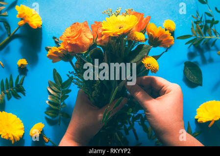 La donna le mani facendo un mazzo di fiori dal vibrante giallo e arancio fiori su una carta blu sullo sfondo. Lay piatto. Foto Stock