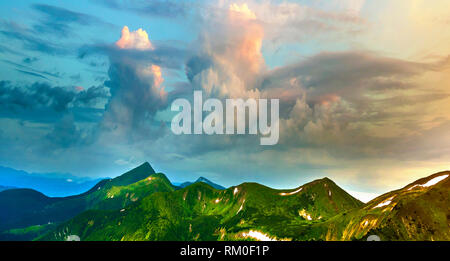 Ampio panorama di illuminata dal sole di mattina valle verde, colline coperte da foreste e distante Misty Mountains con macchie di neve sotto nuvoloso cielo ventoso B Foto Stock