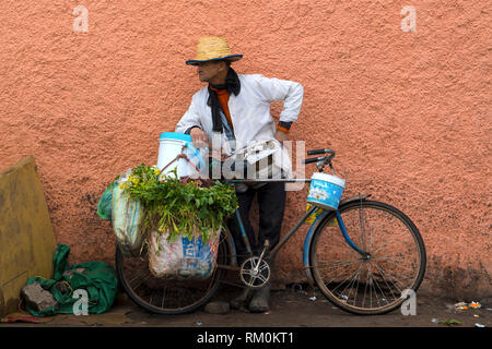 Vecchio burbero uomo vendita di verdure ed erbe del suo vecchio arrugginito bicicletta blu in un cibo di Marrakesh street market in Marocco. Foto Stock