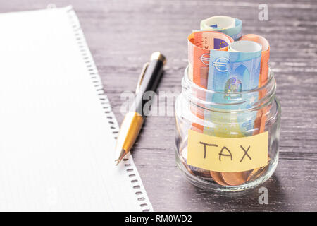 Vaso di vetro con le monete e le banconote in euro con la parola una tassa di tavoli in legno nero. Orologio da polso, penna e carta bianca. Concetto di risparmi finanziari per pagare l Foto Stock