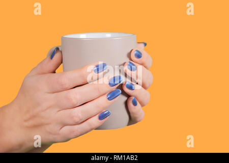 Vista ingrandita di due belle mani femminili con dipinto di blu lucida unghie. Donna che mantiene la tazza isolata su sfondo arancione. Colore orizzontale fotografi Foto Stock