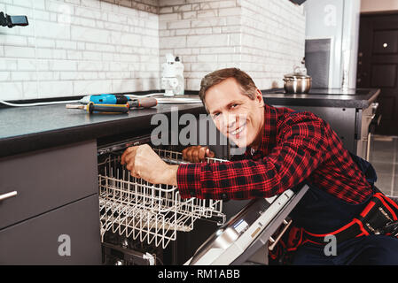 Felice master. Tecnico maschile seduto vicino a lavastoviglie con cacciavite in cucina con strumenti e sorridente Foto Stock