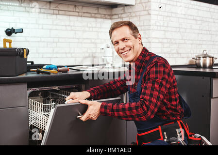 Chiamare il professionista. Tecnico maschile seduto vicino a lavastoviglie con cacciavite in cucina con strumenti e sorridente Foto Stock