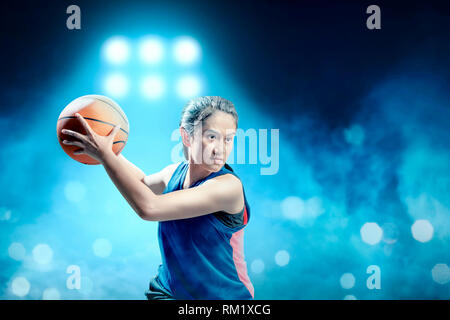 Eccitato ragazza asiatica del giocatore di basket difendendo la palla da avversario sul campo da pallacanestro con faretti blu sullo sfondo Foto Stock