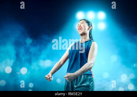 Allegro ragazza asiatica del giocatore di basket in blu sportswear con felicità espressione nel campo da pallacanestro con faretti blu sullo sfondo Foto Stock