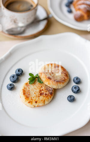 Cheesecake, ai mirtilli freschi e cappuccino. Cottage cheese pancakes o frittelle di cagliata sulla piastra bianca. Sana dieta e colazione all'alba Foto Stock