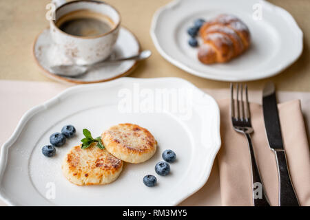 Cheesecake, ai mirtilli freschi e cappuccino. Cottage cheese pancakes o frittelle di cagliata sulla piastra bianca. Sana dieta e colazione all'alba Foto Stock