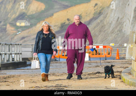 West Bay, Dorset, Regno Unito. Il 13 febbraio 2019. Regno Unito Meteo. Fumoso sole riscalda cane escursionisti che stanno camminando lungo il lungomare di West Bay nel Dorset dopo una fredda per iniziare la giornata. Le temperature sono impostate a salire come venti da sud dai tropici sono impostati a soffiare di tutta la contea nel giro di un paio di giorni portando temperature sopra la media. Credito Foto: Graham Hunt/Alamy Live News Foto Stock