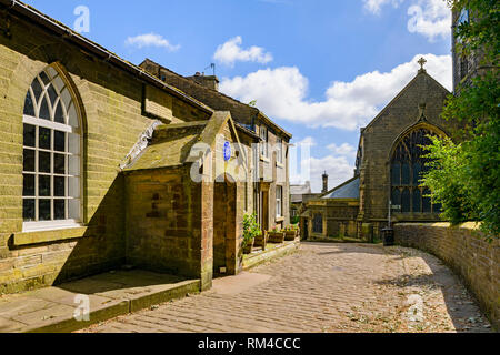 Esterno del soleggiato storica vecchia scuola (Camera Brontes insegnato qui) acciottolate lane & San Michele e Tutti gli Angeli Chiesa - Haworth, West Yorkshire, Inghilterra, Regno Unito Foto Stock
