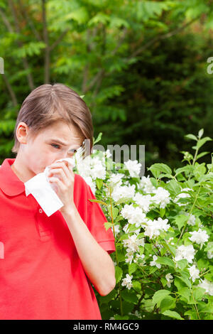 Ragazzo adolescente con influenza stagionale soffia il naso su un tessuto in un giardino di Primavera - stagione concetto di infezione Foto Stock