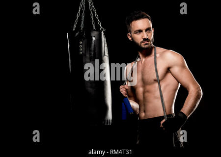 Bello il boxer in piedi con salto con la corda nei pressi di sacco da boxe isolato su nero Foto Stock