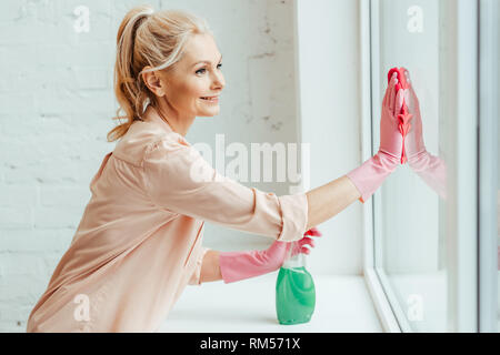 Sorridente donna senior in rosa guanti finestra di pulizia con uno straccio e spray Foto Stock