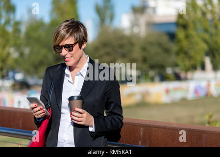 Vista frontale di elegante, sorridente business donna indossando occhiali da sole appoggiato su di una recinzione metallica su un ponte mentre si utilizza il telefono cellulare tenendo un take away Foto Stock