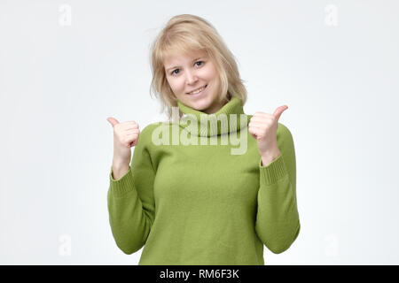 Felice giovane femmina caucasica indossa un maglione verde facendo pollice in alto segno e sorridendo allegramente Foto Stock