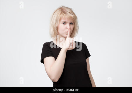Ovely femmina bionda chiede di mantenere segrete le informazioni riservate, vestita di nero per T-shirt Foto Stock