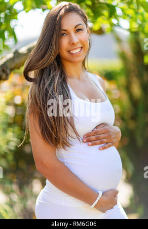 Sorridente donna incinta in piedi in giardino tenendo le mani sulla pancia Foto Stock