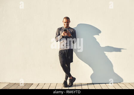 Il relax dopo l'allenamento. A piena lunghezza Ritratto di Allegro atleta africana prendendo una pausa durante gli allenamenti e per chattare con gli amici mentre in piedi contro il muro bianco. Allenamento mattutino. Sport concetto di motivazione. Concetto di fitness Foto Stock