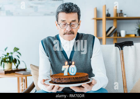 Triste senior uomo con torta di compleanno mentre è seduto su un divano in soggiorno Foto Stock