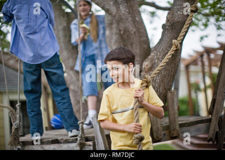 Sorridente ragazzo giovane azienda in una swing corda durante la riproduzione con gli amici nel cortile. Foto Stock