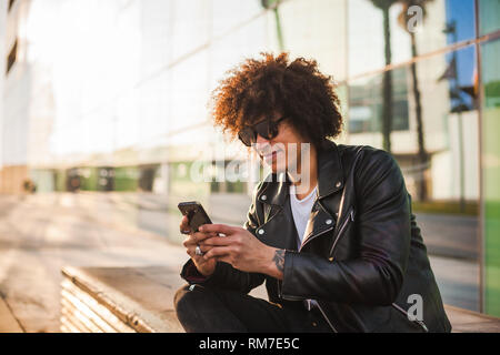 Un bel uomo nero con moderni di acconciatura afro seduti rilassati in strada utilizzando uno smart phone Foto Stock