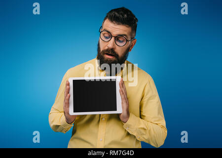 Interessati uomo barbuto guardando a tavoletta digitale con schermo vuoto, isolato su blu Foto Stock