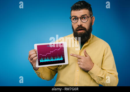 Scioccato uomo barbuto puntando alla tavoletta digitale con tariding app, isolato su blu Foto Stock