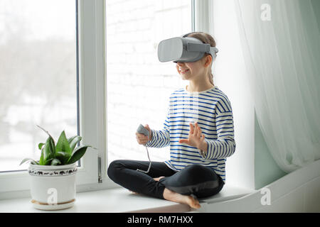 Un bambino con una realtà virtuale auricolare è seduta al tavolo al chiuso in casa. Una ragazza ad esplorare il mondo di realtà virtuale attraverso video e giochi. Foto Stock