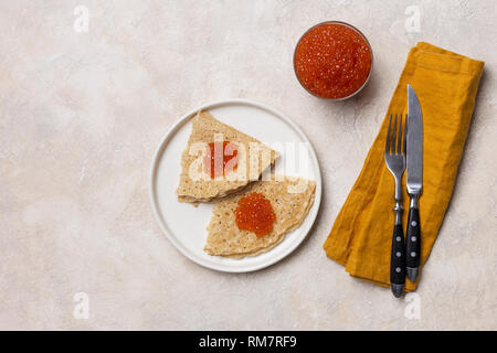 Frittelle di festa con caviale rosso su bianco con piastra forcella, coltello e igienico, un vasetto di caviale, su sfondo bianco. Concetto di cibo festoso. Vista da ab Foto Stock