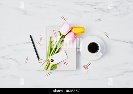 Scrivania da ufficio con tazza di caffè, fermagli per carta, matita, rosa e tulipani gialli con tag per nome o il messaggio sul diario rosa su marmo bianco dello sfondo. Appartamento Foto Stock