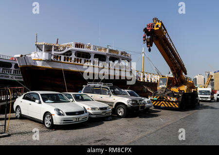 Una flotta di veicoli in attesa di essere ospitati da come gru per essere caricato su un oceano andando Dhow di legno ( barca araba) essendo caricato con carico sul Dubai C Foto Stock