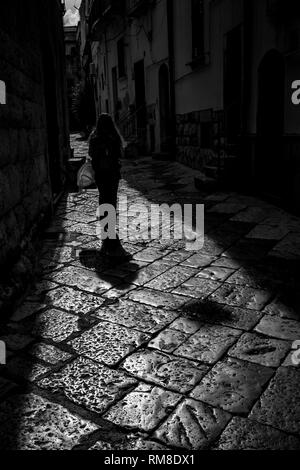 Sun-pietra illuminata strada stretta con una bella ragazza camminare silhouette di Altamura, Puglia, Italia. In bianco e nero ad alto contrasto fotografia presa contro la luce del sole in estate assolato pomeriggio Foto Stock