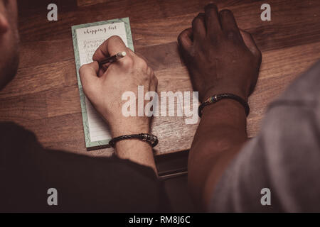 Un interracial gay giovane facendo una lista della spesa per i generi alimentari su una cucina in legno top a casa. Foto Stock