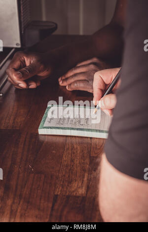 Un interracial gay giovane facendo una lista della spesa per i generi alimentari su una cucina in legno top a casa. Foto Stock