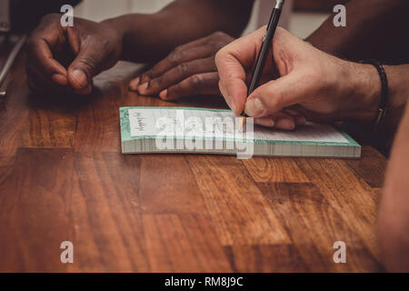 Un interracial gay giovane facendo una lista della spesa per i generi alimentari su una cucina in legno top a casa. Foto Stock