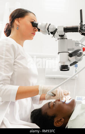 Giovane donna dentista il trattamento di canali radicolari mediante microscopio nella clinica dentale. Uomo paziente sdraiato sulla poltrona del dentista con la bocca aperta. Medicina Foto Stock