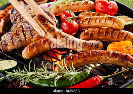 Estate barbecue con sfrigolanti salsicce e le bistecche alla fiorentina la cottura su una griglia su un fuoco con un fresco e peperoni, peperoncino, pomodoro e zucchine condito Foto Stock